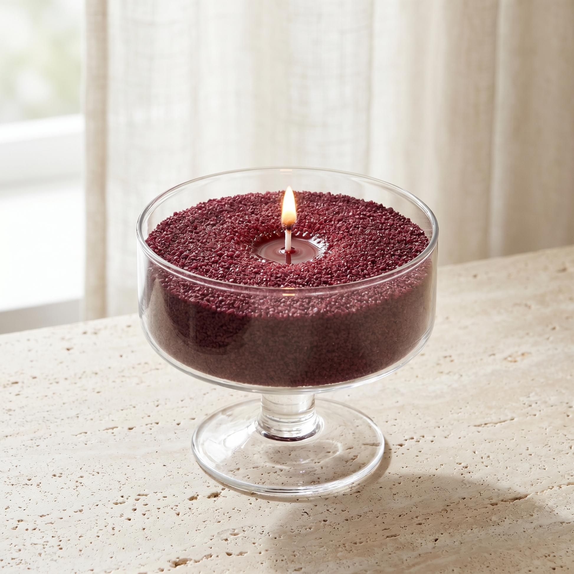 Drunken Pash wine-red sand wax candle, lit, in a footed glass on travertine